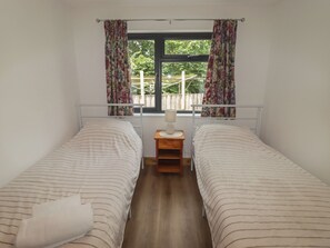 A bedroom with two beds, a window with curtains, a bedside table with a lamp, and a view of greenery outside. - Brookview Goldfinch (Haverfordwest)