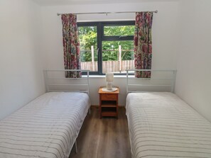 A room with two single beds, a window with floral curtains, a bedside table with a lamp, and a view of greenery outside. - Brookview Goldfinch (Haverfordwest)