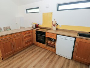 A compact kitchen with wooden cabinets, a built-in oven, a dishwasher, and a small window above the sink. - Brookview Goldfinch (Haverfordwest)