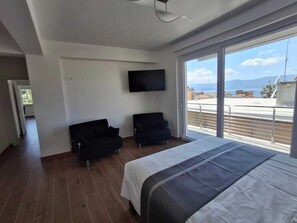 Una camera d'albergo moderna con un letto grande, due poltrone nere, una TV a schermo piatto montata a parete e un balcone con vista sul mare.