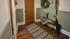 Un pasillo con una puerta de madera, un banco con cojín verde, un adorno colgante y un tapete a rayas.