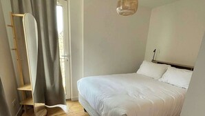 Et soveværelse med en seng, en hængende lampe, trægulv og et vindue med gardiner.