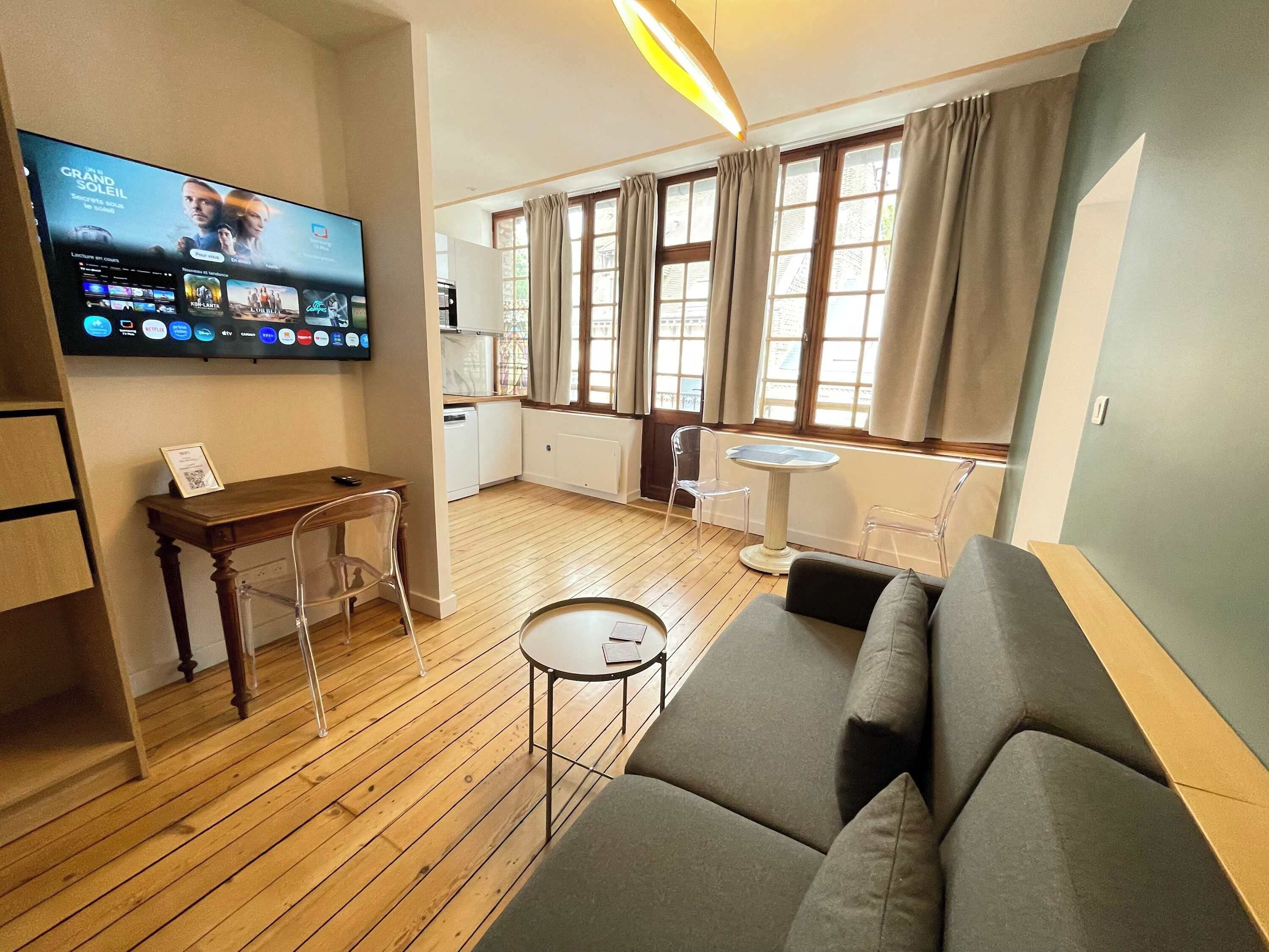 Una sala de estar con un sofá, una mesita, un televisor montado en la pared y una cocina visible a través de una puerta abierta.