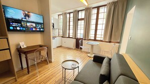 En stue med sofa, et lille bord, et fjernsyn ophængt på væggen og et køkkenområde, der kan ses gennem en åben dør.