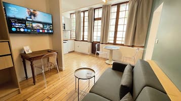 Una sala de estar con un sofá, una mesita, un televisor montado en la pared y una cocina visible a través de una puerta abierta.
