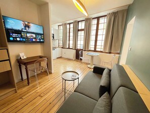 Ein Wohnzimmer mit einer Couch, einem kleinen Tisch, einem an der Wand befestigten Fernseher und einem durch eine offene Tür einsehbaren Küchenbereich.