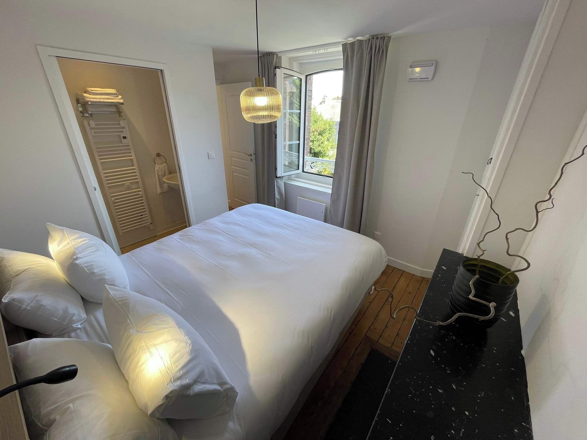 Een slaapkamer met een bed, nachtkastje, hanglamp, raam met gordijnen en een deur naar een badkamer.