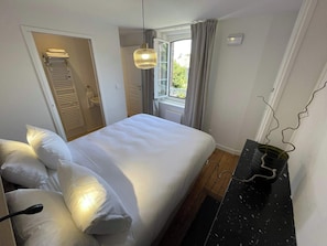 Quarto com cama, mesa de cabeceira, luminária pendente, janela com cortinas e porta para um banheiro.