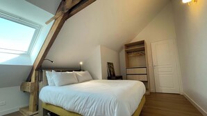 A bedroom with a sloped ceiling, a bed with white bedding, a bedside lamp, a wooden headboard, a closet with hanging space, and a window. - Spacious Duplex • 2 Suites • Space & Privacy • Fully Equipped • Amiens Centre • 5-min Walk to Station • Parking on Site (Amiens)