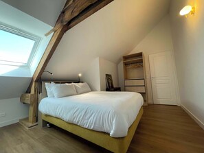 Ein Schlafzimmer mit schrägverdachter Decke, einem Bett mit weißer Bettwäsche, einer Nachttischlampe, einem Holz-Kopfteil, einem Kleiderschrank mit Ablagefläche und einem Fenster.