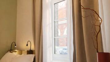 Une chambre d’hôtel avec un lit, une table dressée pour le repas, une fenêtre avec des rideaux et un vase décoré d’une branche.