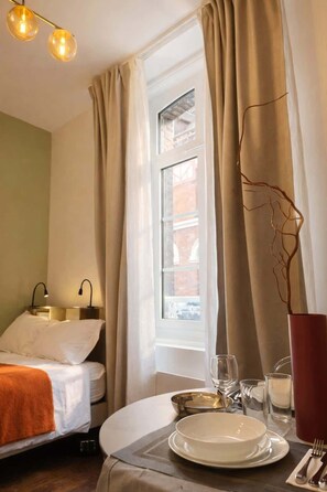 Ein Hotelzimmer mit einem Bett, einem Esstisch, einem fenster mit Vorhängen und einer Vase mit einer Zweigdekoration.