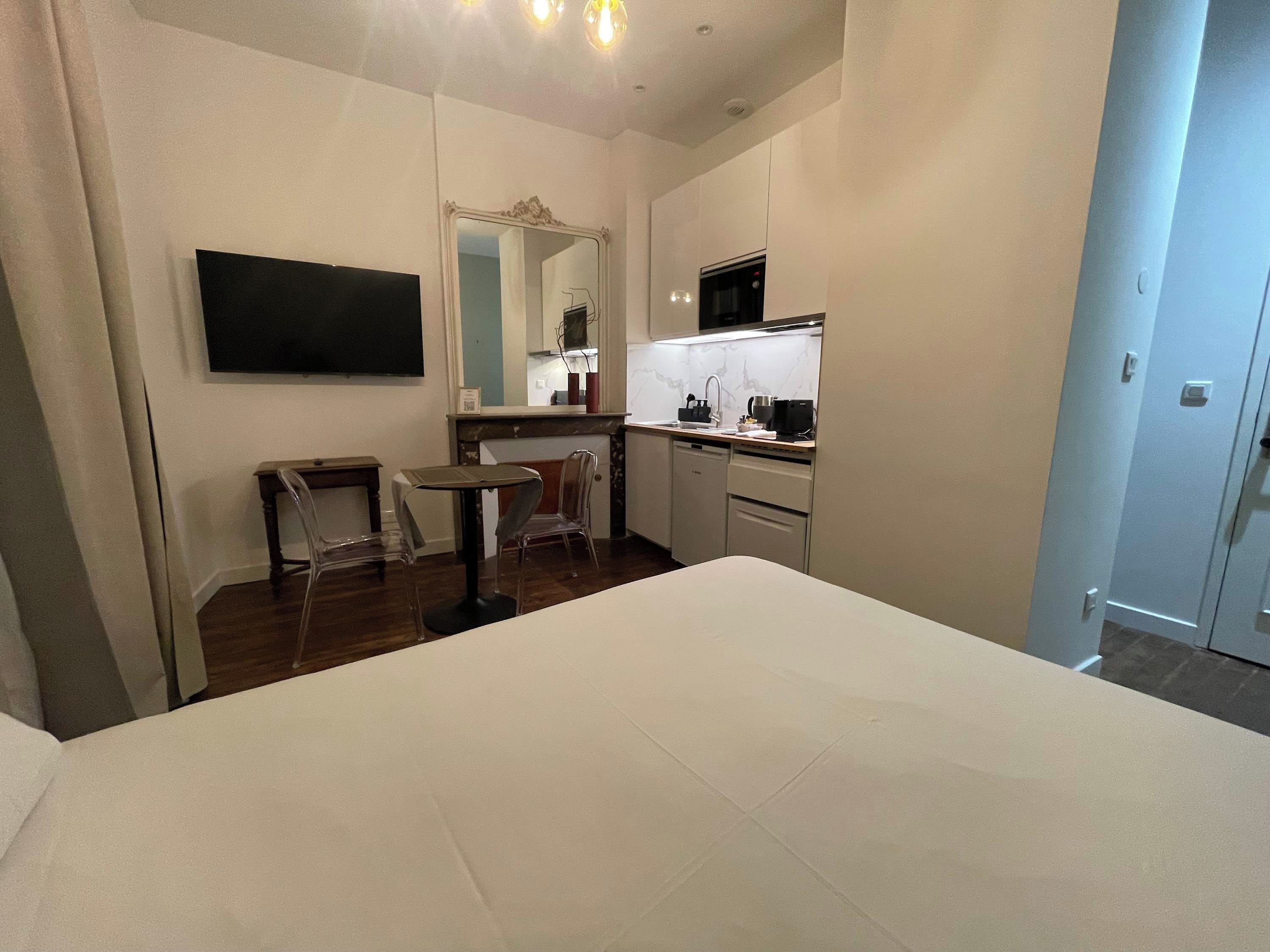 Une chambre d’hôtel comprenant un lit, une petite table, des chaises, un téléviseur, un coin cuisine avec un four à micro-ondes et un grand miroir.