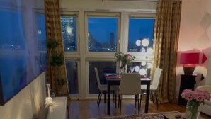 Una sala de estar moderna con un televisor de pantalla plana, un comedor con sillas y una mesa, y ventanas amplias con vista a la ciudad.