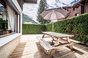 Une terrasse en bois avec une table de pique-nique et un parasol, entourée d’une haie et offrant une vue sur la montagne.