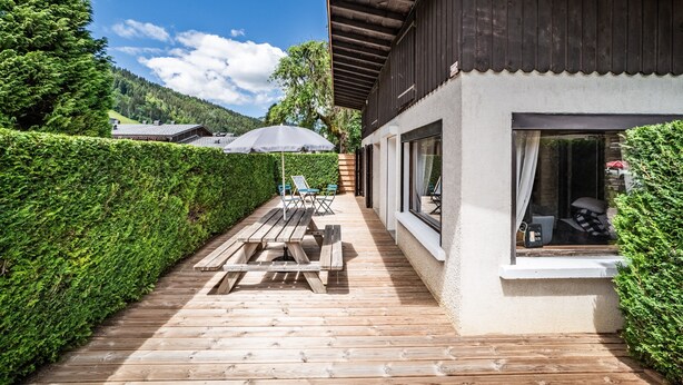 Un molo in legno con tavoli da picnic e ombrelloni, un edificio con ampie finestre e un vialetto delimitato da siepi.