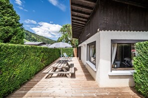 Une terrasse en bois avec des tables de pique-nique et des parasols, un bâtiment doté de grandes fenêtres et un sentier bordé de haies.