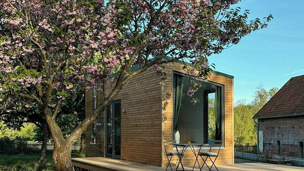 A modern wooden cabin with large windows, a deck, and a tree with pink blossoms. - Modern tiny house with private lake access (Walsrode)