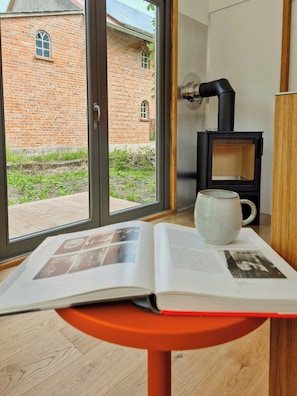 Ein Zimmer mit einem großen Fenster, einem Buch auf einem kleinen Tisch, einer weißen Tasse und einem schwarzen Kaminofen.