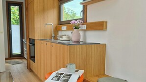 A modern kitchen with wooden cabinets, a built-in oven, a window, and a small table with a vase of flowers. - Modern tiny house with private lake access (Walsrode)