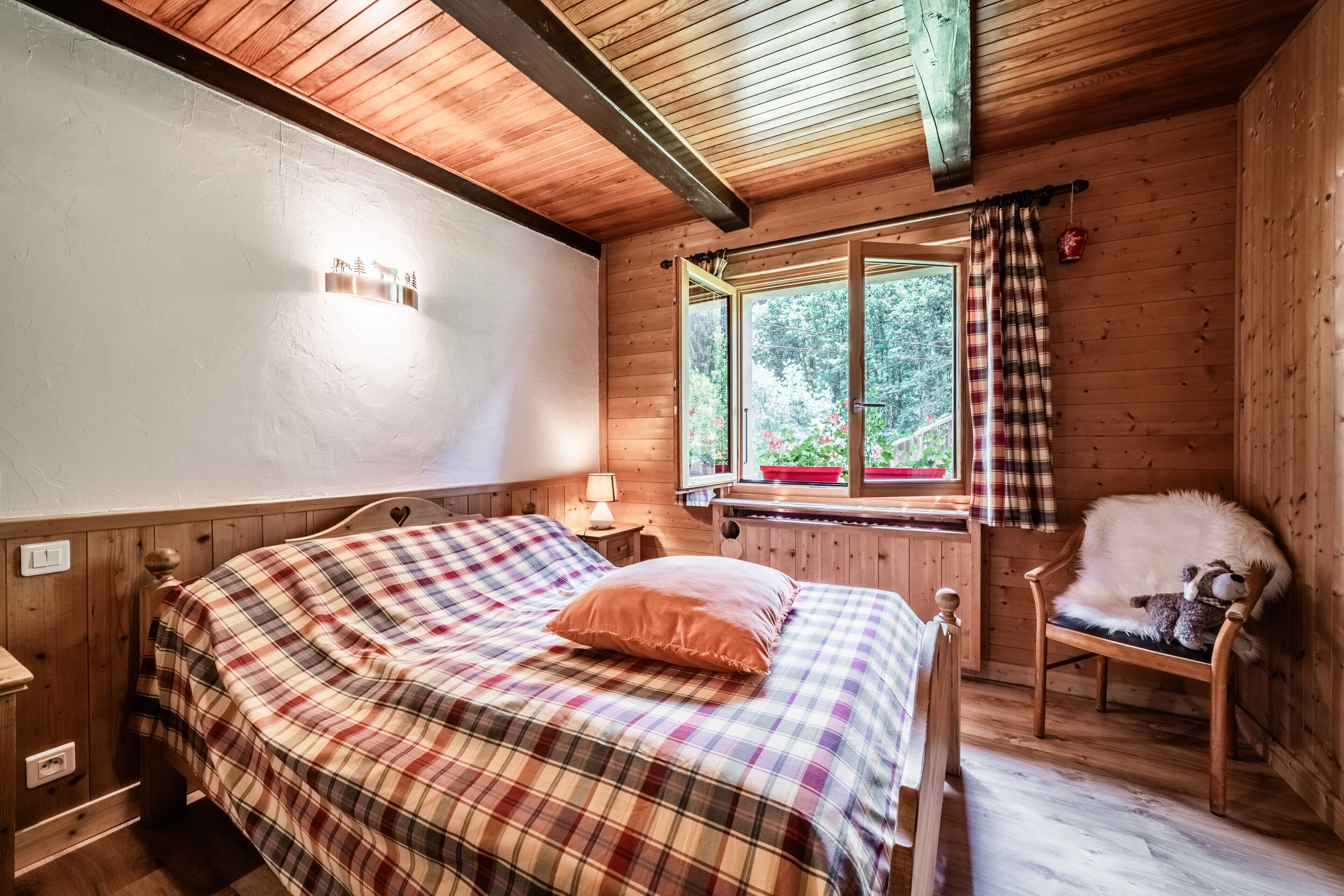 Un dormitorio de cabaña de madera con una cama, una ventana con cortinas, una silla y una lámpara.