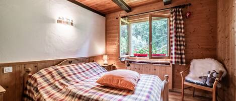 Un dormitorio de cabaña de madera con una cama, una ventana con cortinas, una silla y una lámpara.
