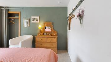 Un dormitorio con cama, cómoda, silla, perchero y una pared con un cuadro y una lámpara.