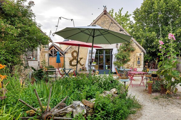 A rustic outdoor seating area with tables and chairs, a large umbrella, and a building with a distinctive roof.