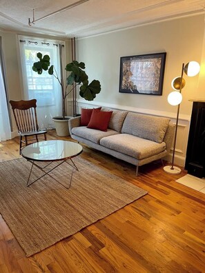 A living room with a sofa, a coffee table, a chair, a large plant, and a painting on the wall. - Brooklyn Garden Apartment (Brooklyn)