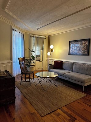 A living room with a sofa, a glass coffee table, a wooden chair, a rug, and a painting on the wall. - Brooklyn Garden Apartment (Brooklyn)