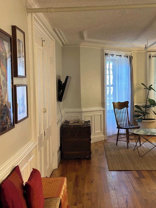 Ein Zimmer mit Holzboden, einem Sessel, einem Tisch, einem an der Wand befestigten Fernseher und einer Tür mit weißem Rahmen.