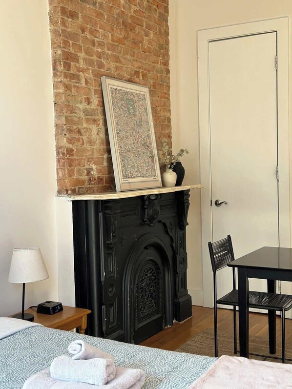 Una habitación con chimenea de ladrillo, un cuadro enmarcado, una puerta blanca, una mesa negra con una silla y una cama con colcha estampada.
