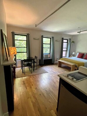 Ein Zimmer mit Holzboden, einem Bett mit roten und grauen Kissen, einem Schreibtisch mit Lampe sowie zwei mit Vorhängen versehenen Fenstern.