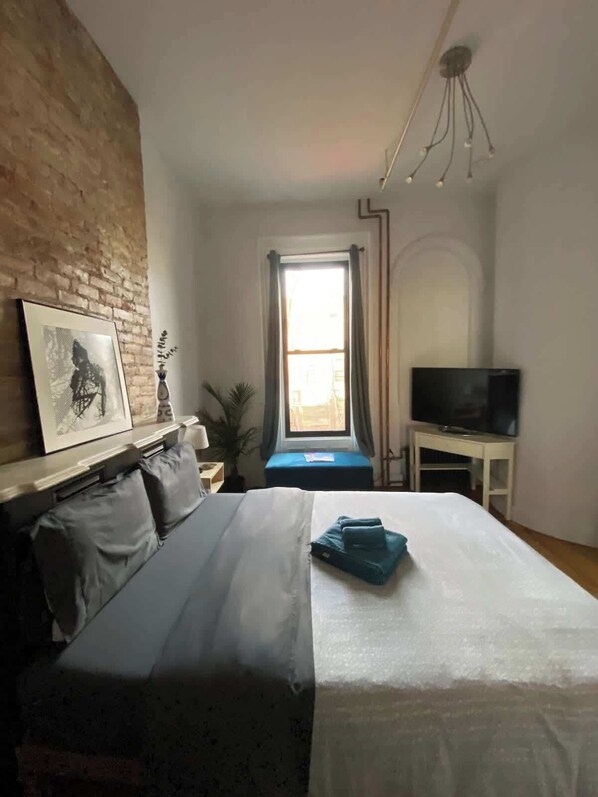Quarto com cama, televisão, janela com cortinas, parede de tijolos e um quadro na parede.