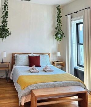 Een bed met een houten hoofdeinde, twee nachtkastjes, een bankje en een raam met gordijnen.