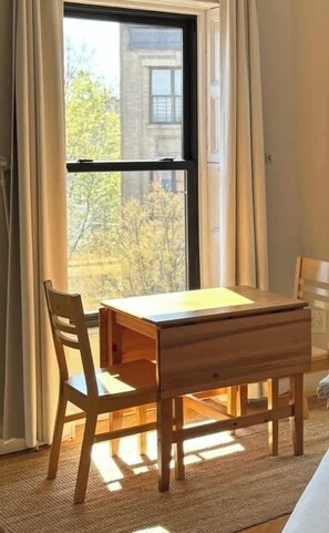 Ein Zimmer mit Holztisch und Stühlen, einem großen Fenster mit Vorhängen und Blick auf Bäume und ein Gebäude.