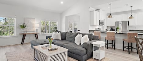 A spacious living area with a grey sofa, white coffee table, and a kitchen with white cabinets and stainless steel appliances.