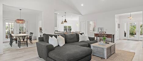A spacious living area with a grey sectional sofa, a dining table with chairs, and a kitchen in the background.