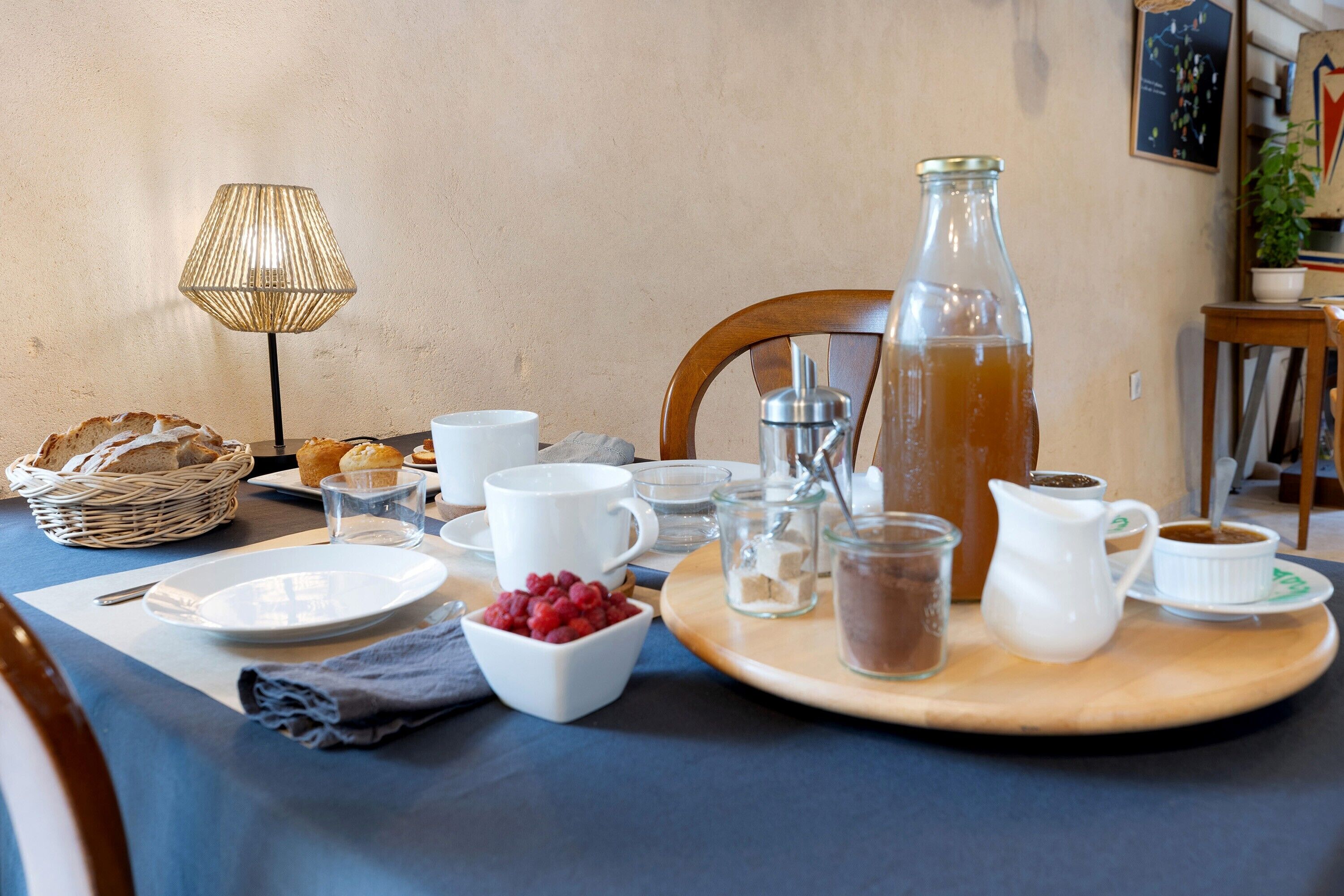 Une table dressée pour le petit-déjeuner, avec des assiettes, des tasses, un pichet, un panier et une lampe.