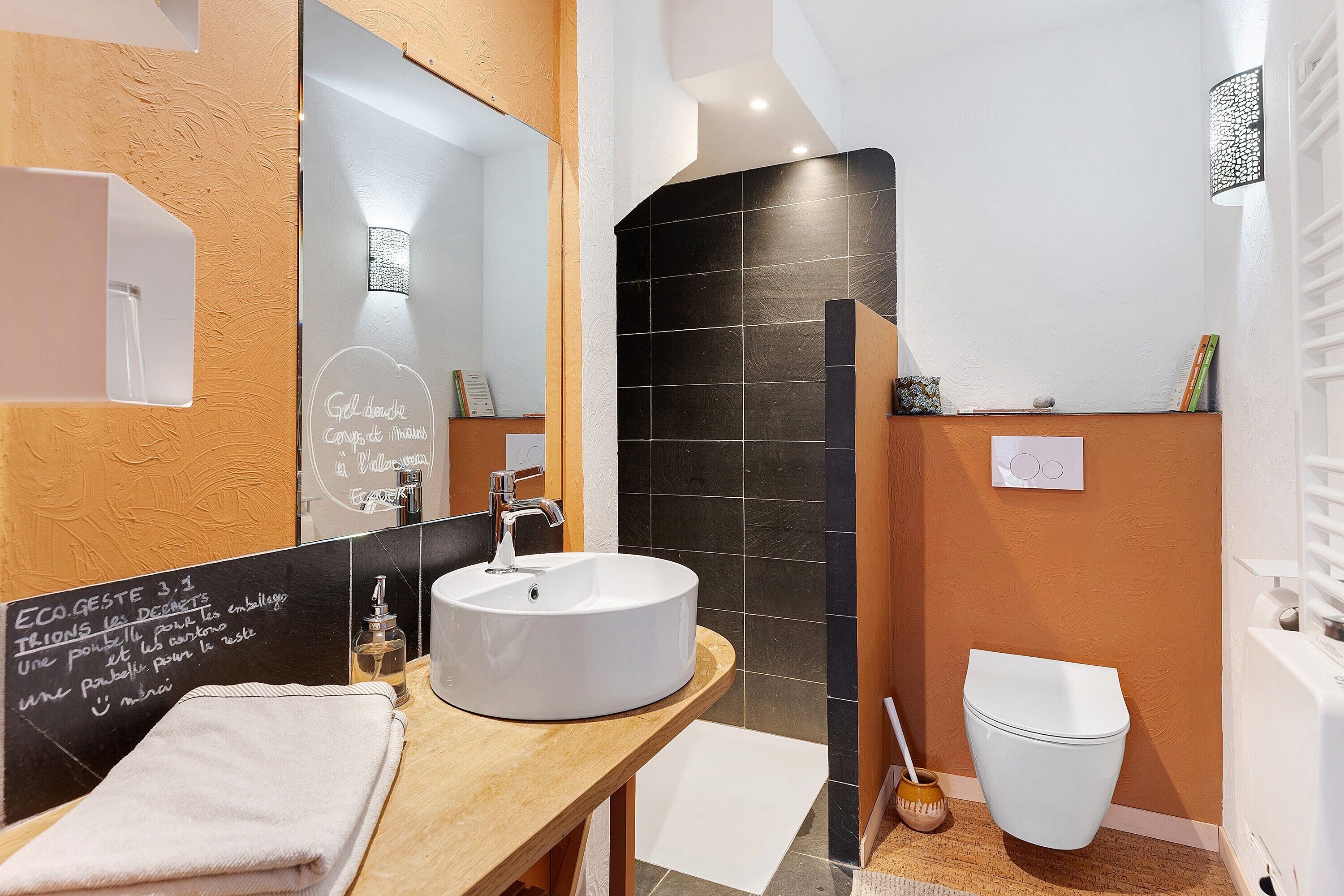 Un baño con lavamanos blanco, espejo, pared de azulejos negros y retrete blanco.
