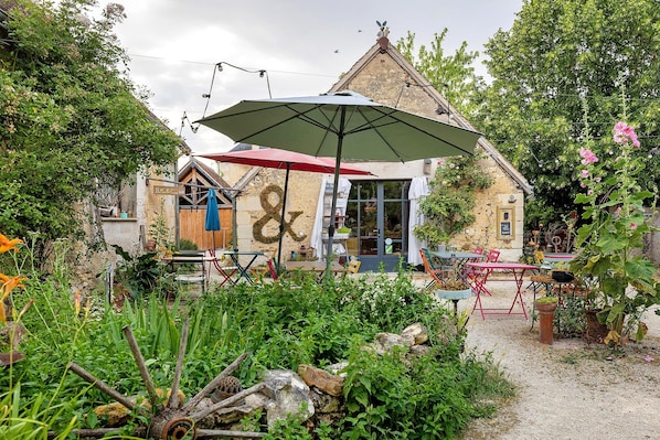 Un espace extérieur aménagé dans un style rustique, avec des tables et des chaises, un grand parasol et un bâtiment doté d’un toit remarquable.
