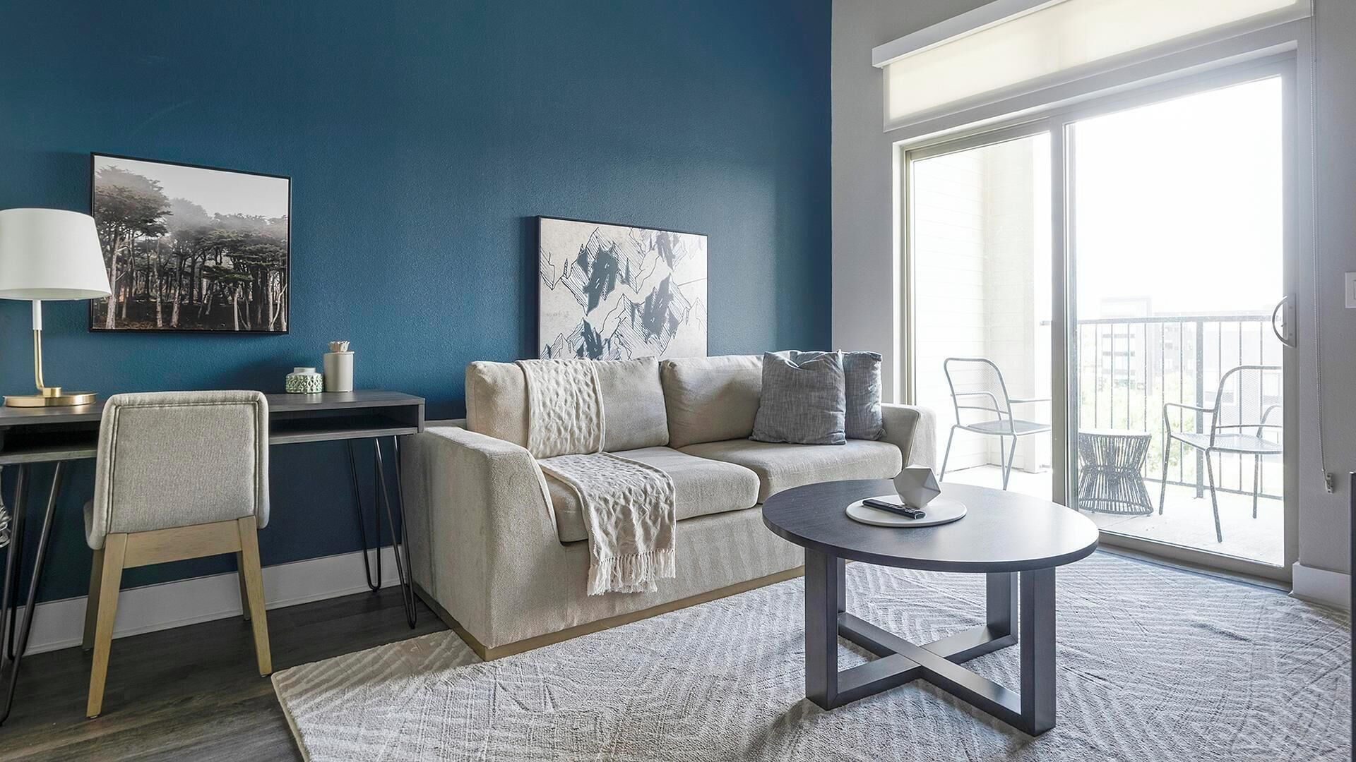A living room with a sofa, a coffee table, a desk, and a chair. There is a balcony with chairs outside.