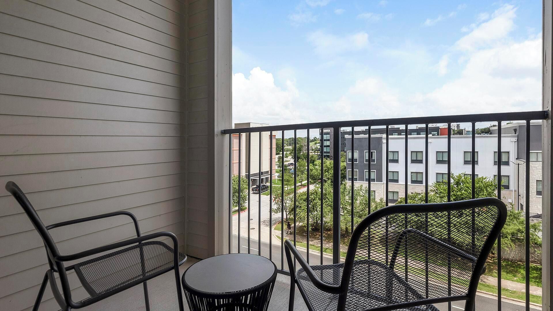 A balcony with two chairs and a table, overlooking a street with buildings and greenery.