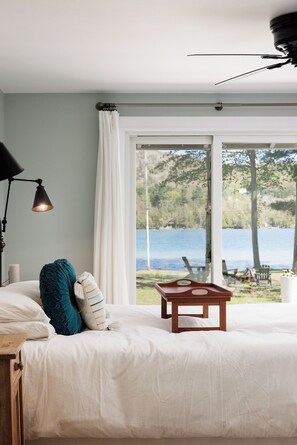 Une chambre Ă coucher comprenant un lit, une table de chevet, une lampe, une fenĂȘtre donnant sur un lac et des arbres, et un ventilateur de plafond.