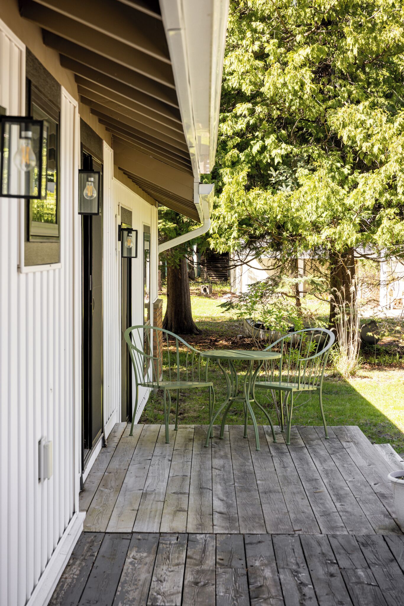 Une terrasse en bois avec une table et des chaises en métal, un bâtiment blanc avec une porte, et un arbre en arrière-plan.