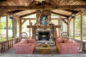 A covered outdoor seating area with a stone fireplace, checkered sofas, and a view of the outdoors.