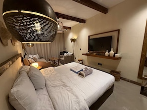 A bedroom with a large bed, a television mounted on the wall, a hanging light fixture, and a wooden ceiling beam.