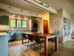 A modern kitchen with a dining area, wooden flooring, and a view of greenery through the windows.