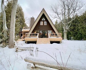 A cabin with a triangular roof, wooden deck, and a staircase leading to the entrance. - Goathaus | Iconic A-Frame with Bay Views (Sister Bay)