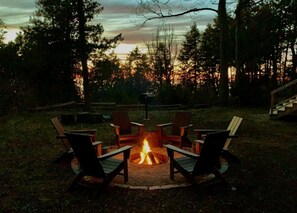A fire pit with four Adirondack chairs arranged around it, set in a grassy outdoor area with trees in the background.